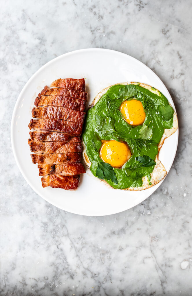 Overhead shot of two fried eggs with salsa verde and a pork chop on a white plate and marbled background