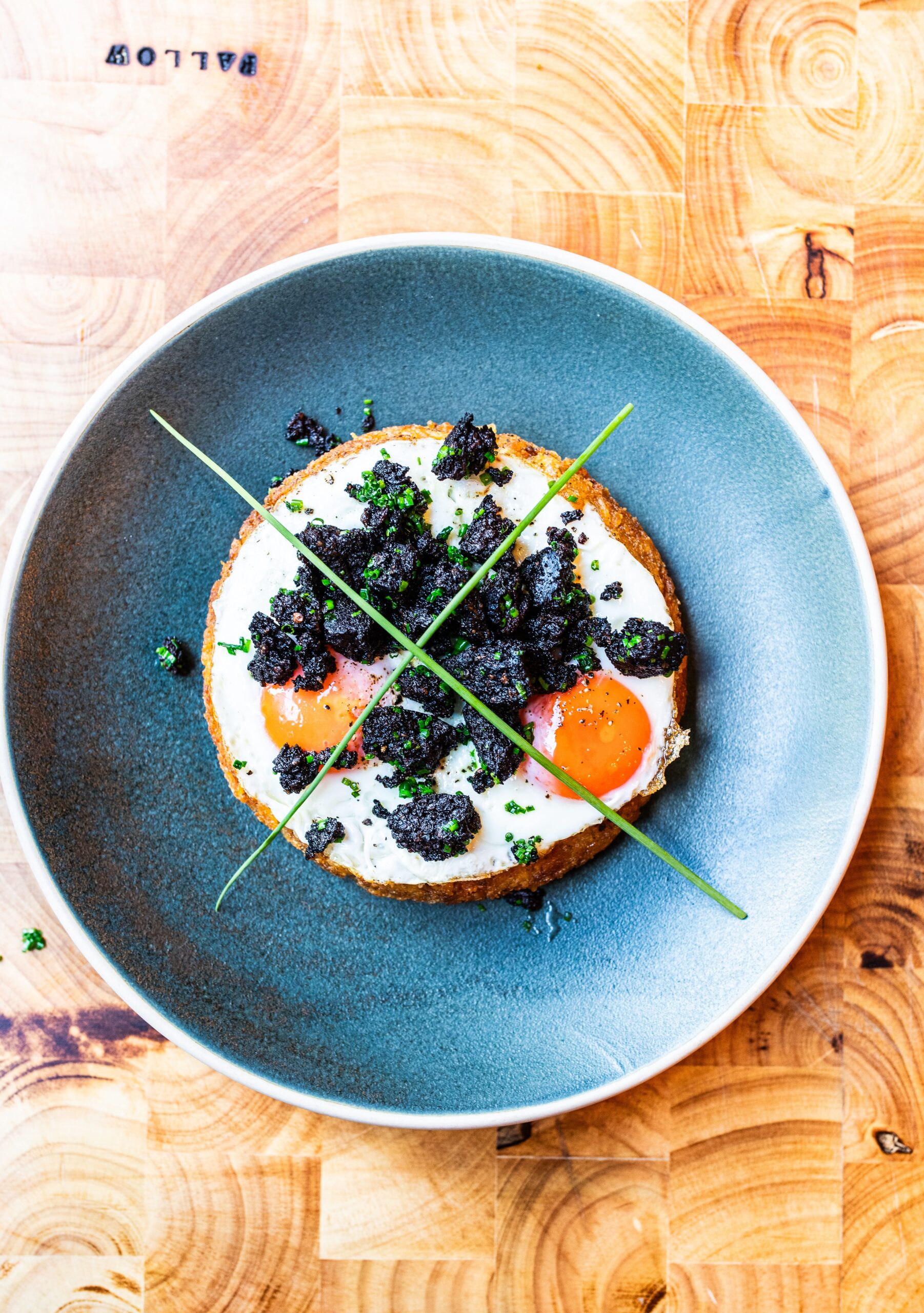 Overhead shot of black pudding rösti with two fried eggs on top. All on a blue plate on a wooden background.