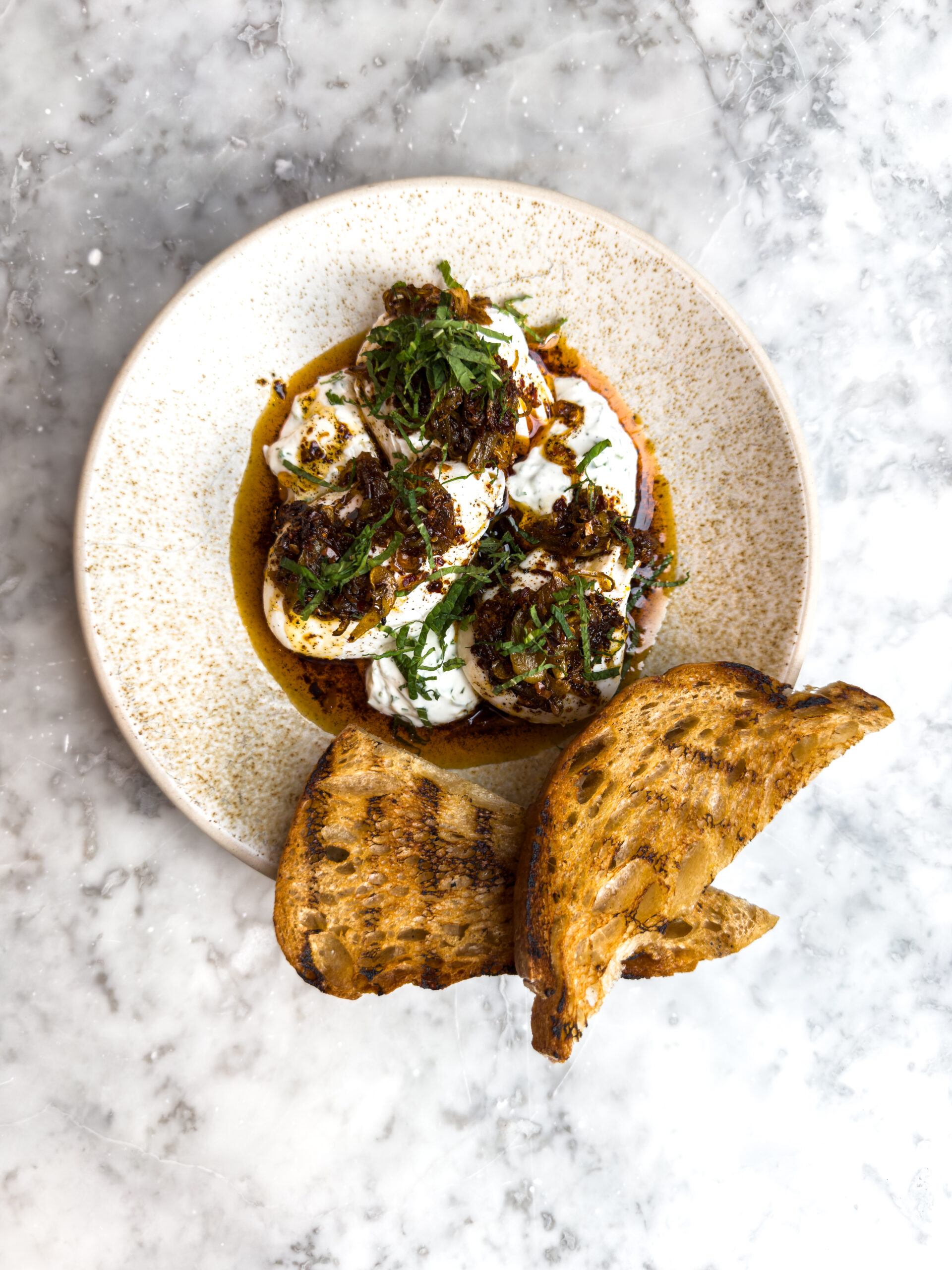 Overhead view of brown butter Turkish eggs in a beige speckled bowl with grilled sourdough toast, all on a marble background