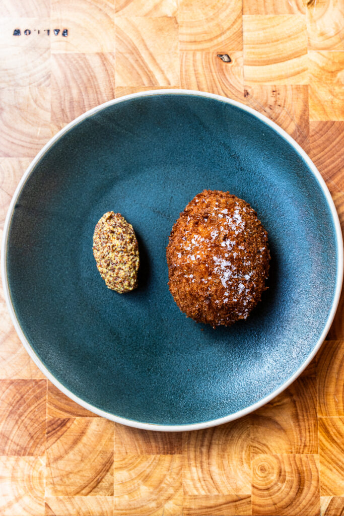 Overhead view of a bubble and squeak scotch egg with wholegrain mustard, all on a blue plate and wooden table top.