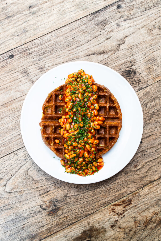 Overhead view of sriracha beans on a homemade potato waffle. All on a white plate and wooden table top.