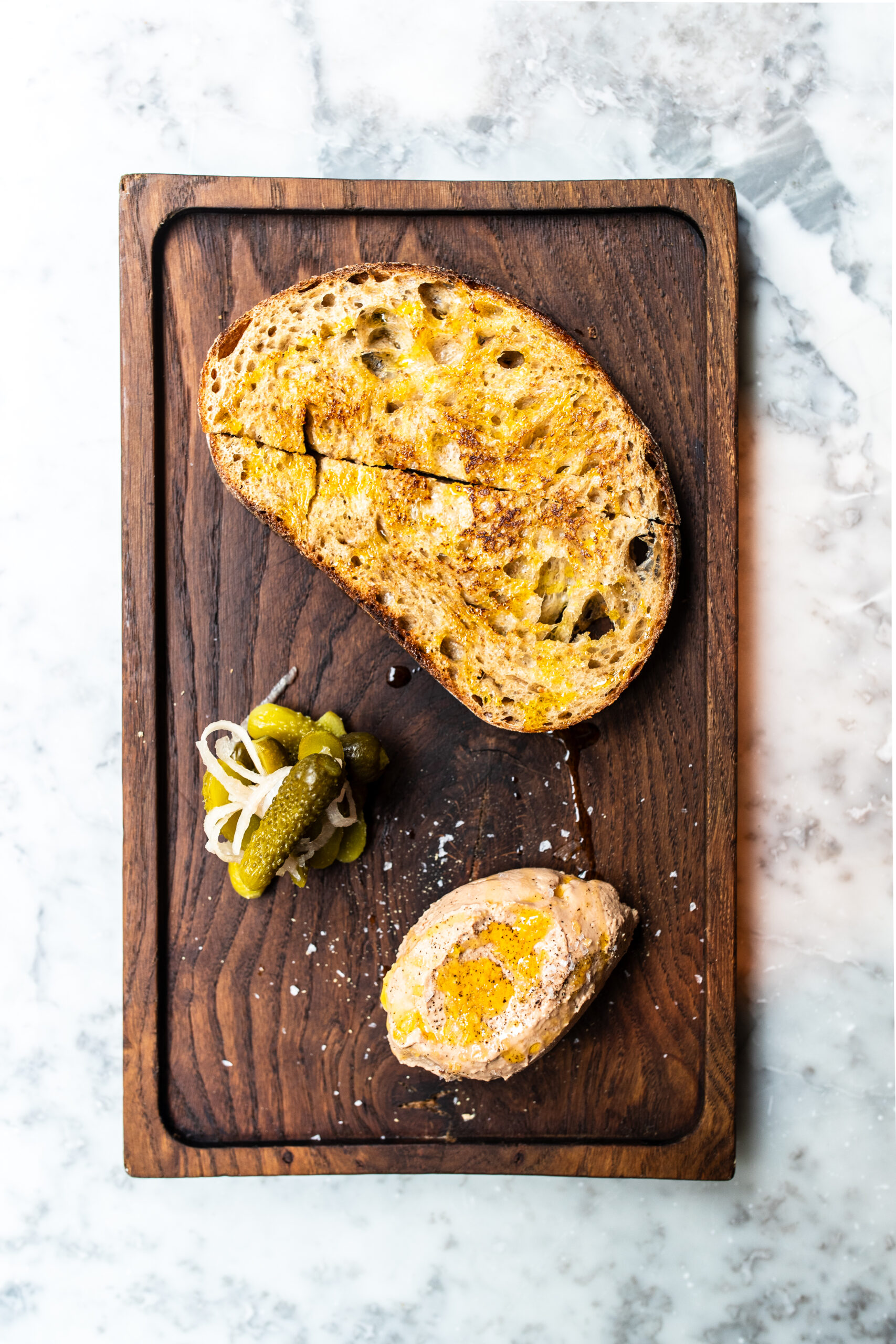 An overhead image of chicken liver parfait and bread on a wooden chopping board on a white marble table.