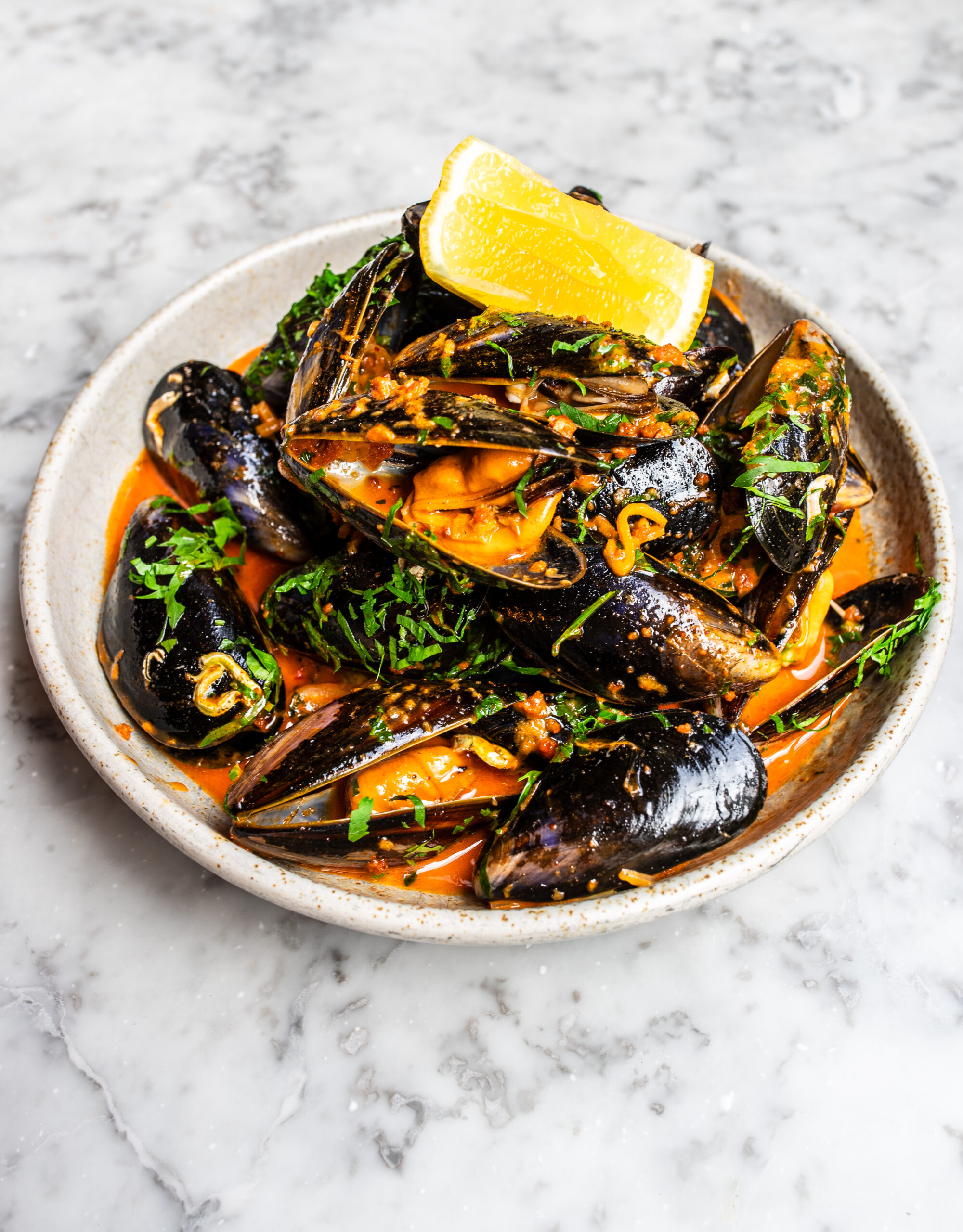 An overhead shot of a white bowl filled with mussels in an 'nduja butter sauce topped with parsley and a wedge of lime on a white marble table