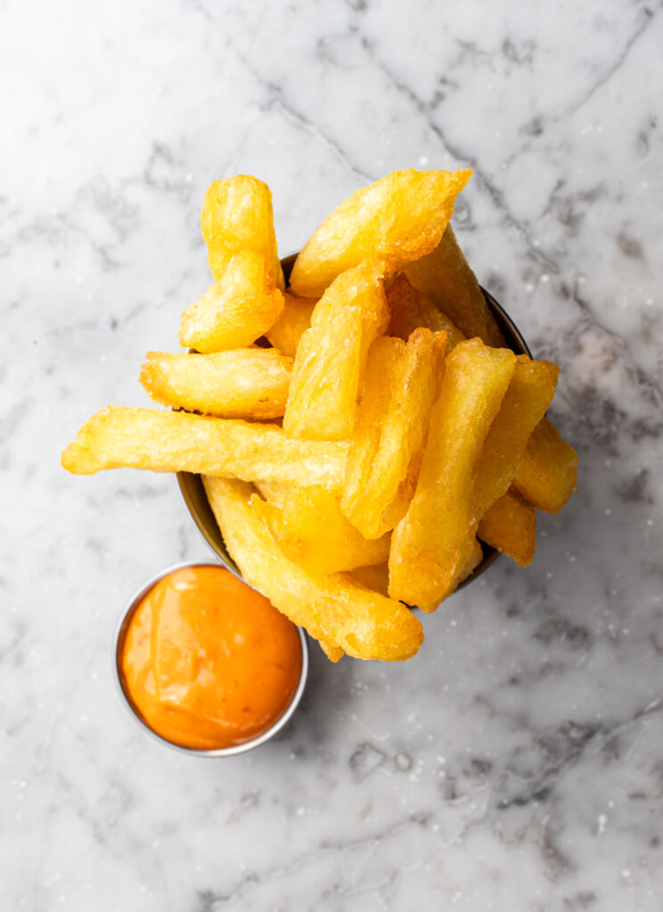An overhead shot of triple-cooked chips with a small pot of sriracha mayonnaise on the side, all on a white marble table
