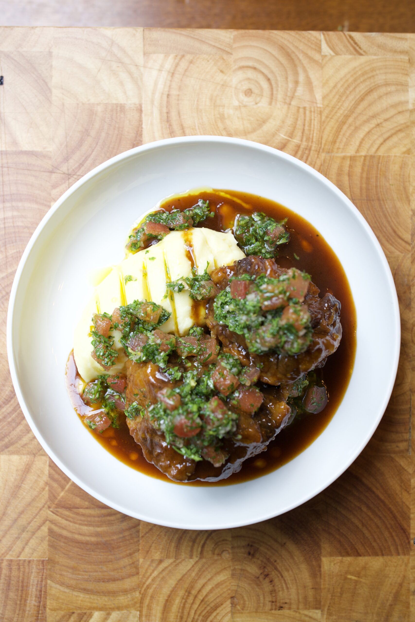 An overhead image of braised oxtail with mashed potatoes and persillade in a white bowl, on a wooden table.