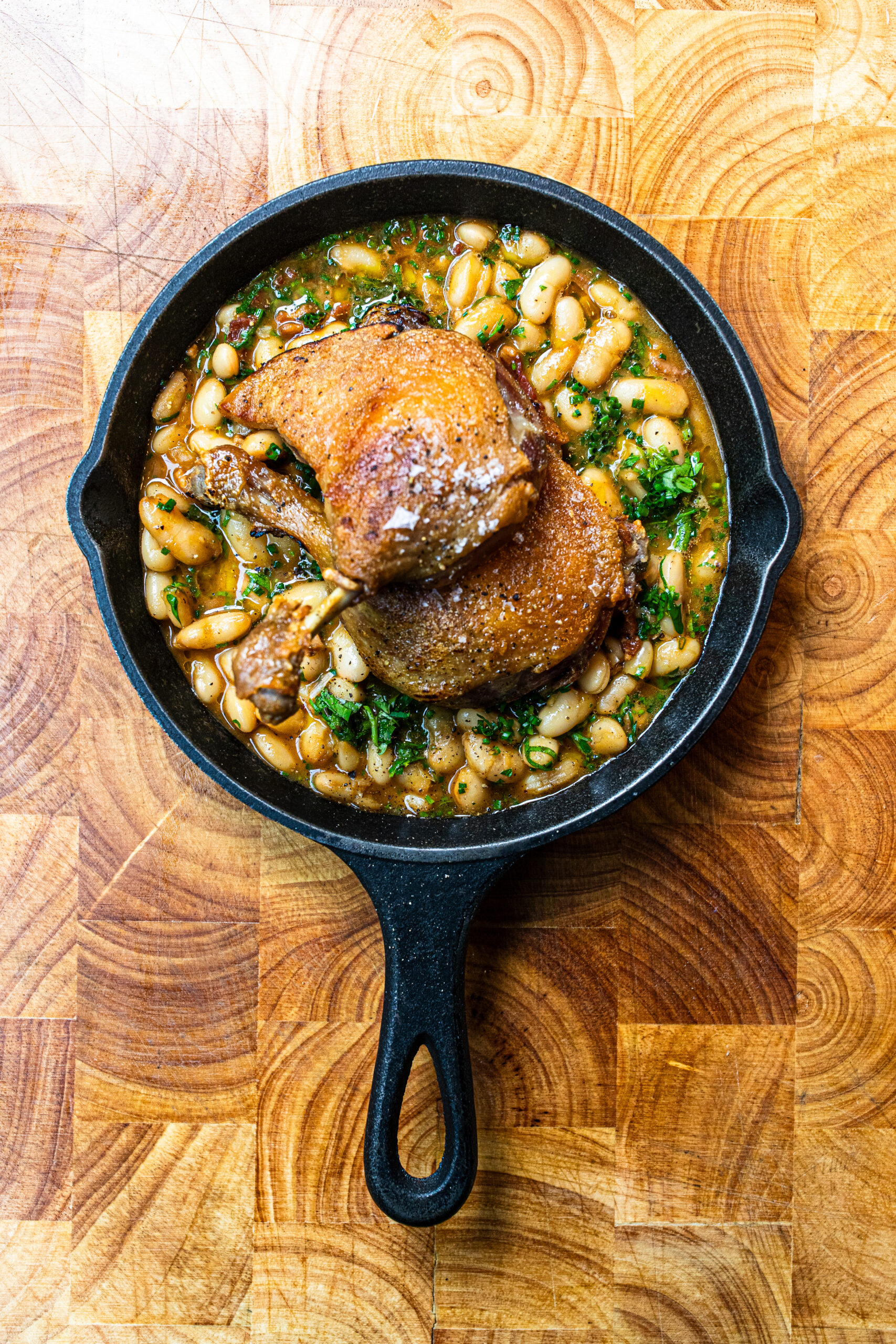 An overhead image of 2 confit duck legs on top of white beans and herbs in a black frying pan on a wooden table.