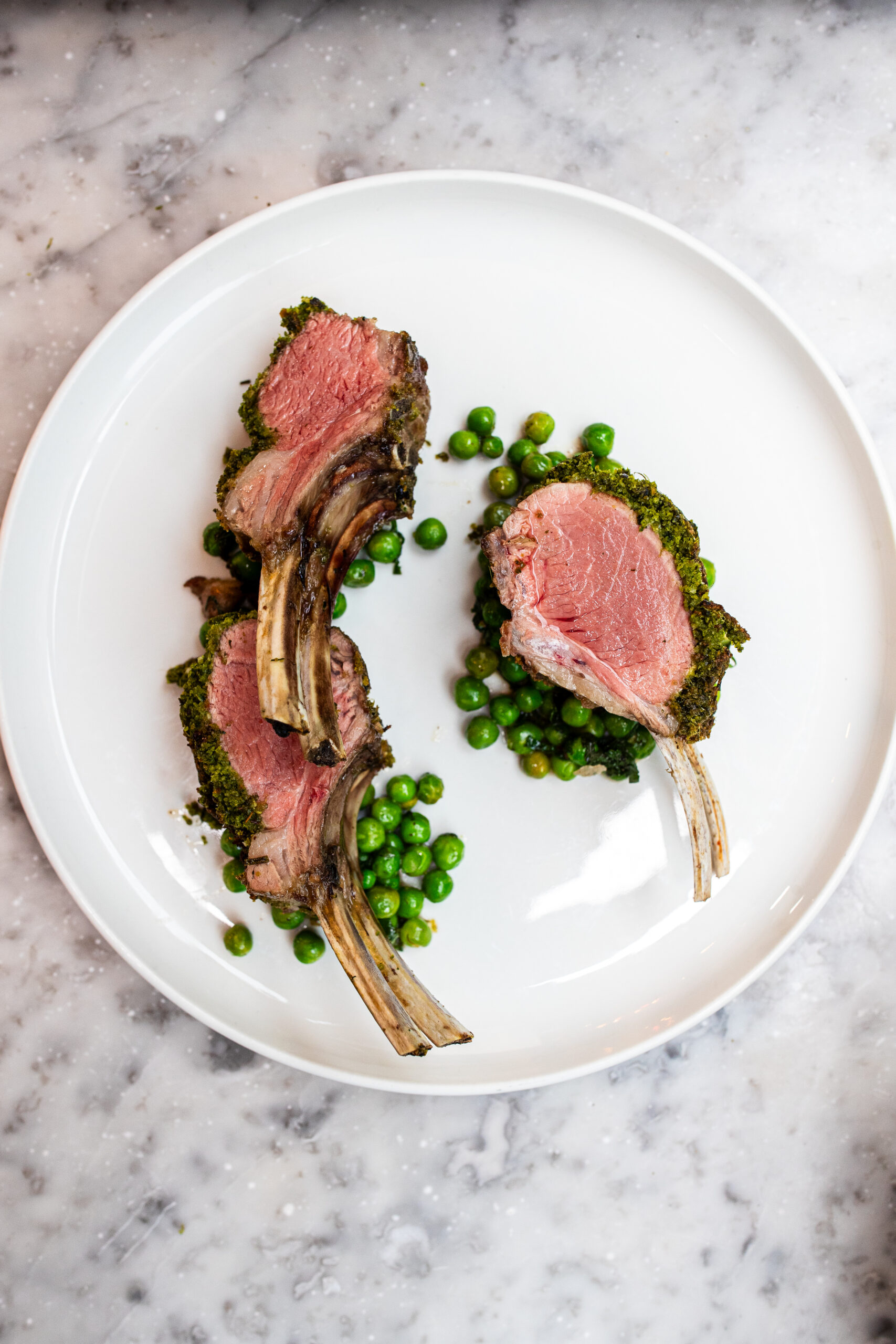 On overhead shot of 3 carved pieces of lamb from a rack of life, on top of peas on a white plate on top of a white marble table.